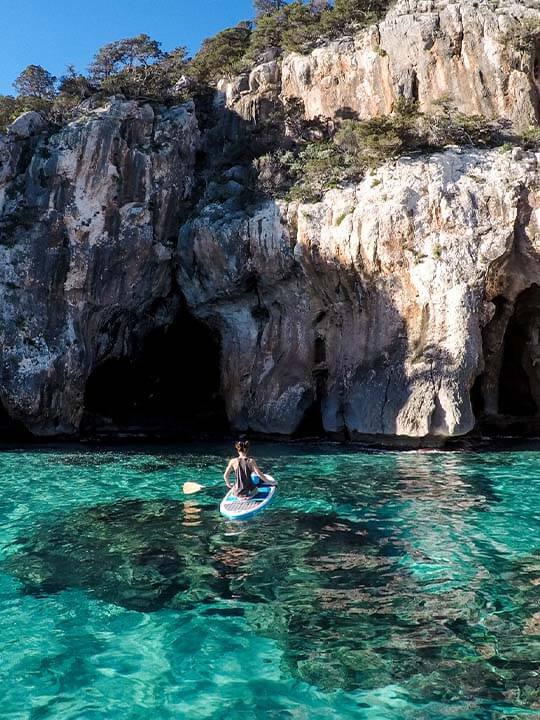 Man Paddle Boarding in Blue Crystal Clear Water With Cliff and Caves Infront of Him